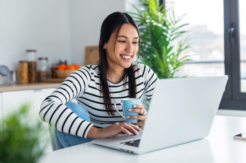 Shot of smiling young woman watching training video with laptop while holding a cup of coffee in the kitchen at home.